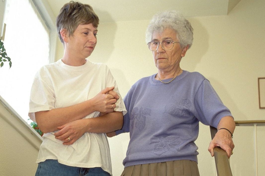 Eine jüngere Frau hilft einer Seniorin, eine Treppe hinabzugehen. Sie hält die ältere Frau am Arm eingehakt fest.