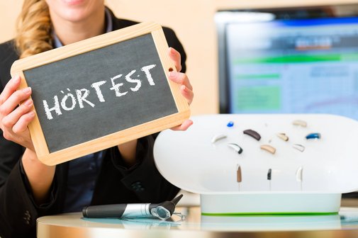 Eine Frau h&auml;lt eine Schiefertafel, auf der das Wort H&ouml;rtest steht. Daneben liegen verschiedene kleine H&ouml;rger&auml;te. 