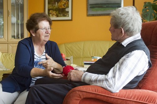Pflege zuhause Eine ältere Frau sitzt ihrem Mann gegenüber. Er hält einen roten Ball in den Händen.