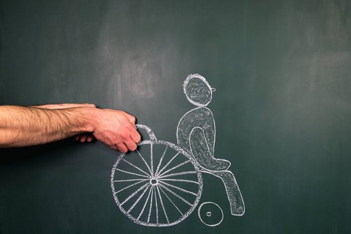 Pflegebedürftigkeit Ein Männchen, das im Rollstuhl sitzt, ist mit weißer Kreide an eine Tafel gezeichnet. Jemand hält seine Hände vor die Zeichung, so dass es aussieht, als würde er den Rolstuhl schieben.