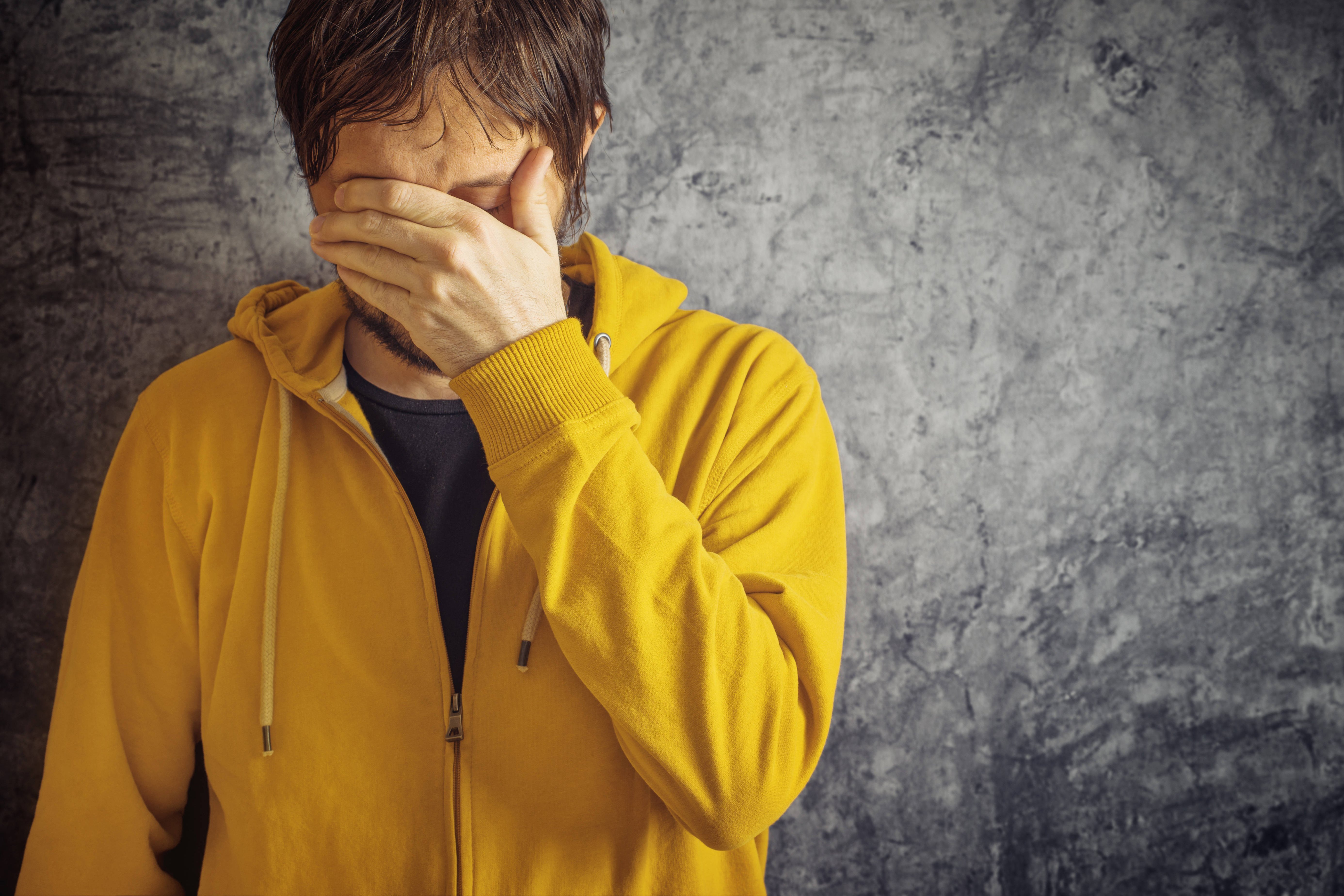 Ein Mann in gelber Sweatjacke steht vor einer grauen Wand, er hält sich eine Hand vor die Augen, senkt den Kopf