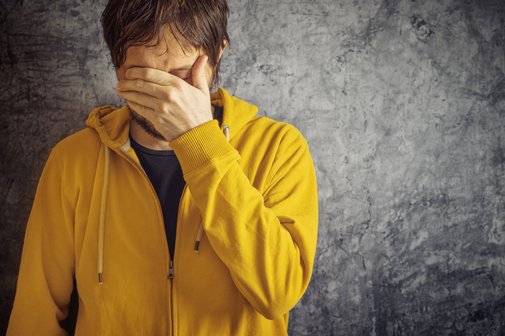 Ein Mann in gelber Sweatjacke steht vor einer grauen Wand, er h&auml;lt sich eine Hand vor die Augen, senkt den Kopf