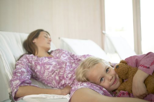 Eine Frau liegt mit geschlossenen Augen auf einer Liege, die aussieht wie eine Liege in einer Gesundheitseinrichtung. Ein kleines Mädchen ist an die Frau gekuschelt, blickt in die Kamera.