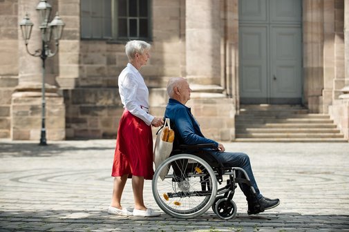 Eine Seniorin schiebt ihren Mann, der im Rollstuhl sitzt, &uuml;ber einen Platz
