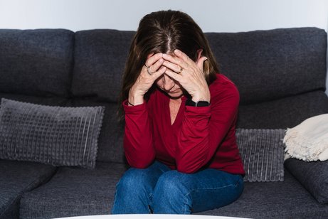 Erschöpfte Frau Eine ältere Frau sitzt auf einem Sofa, hat den Kopf gesenkt und hält sich die Hände vor das Gesicht. Sie sieht erschöpft oder traurig aus.