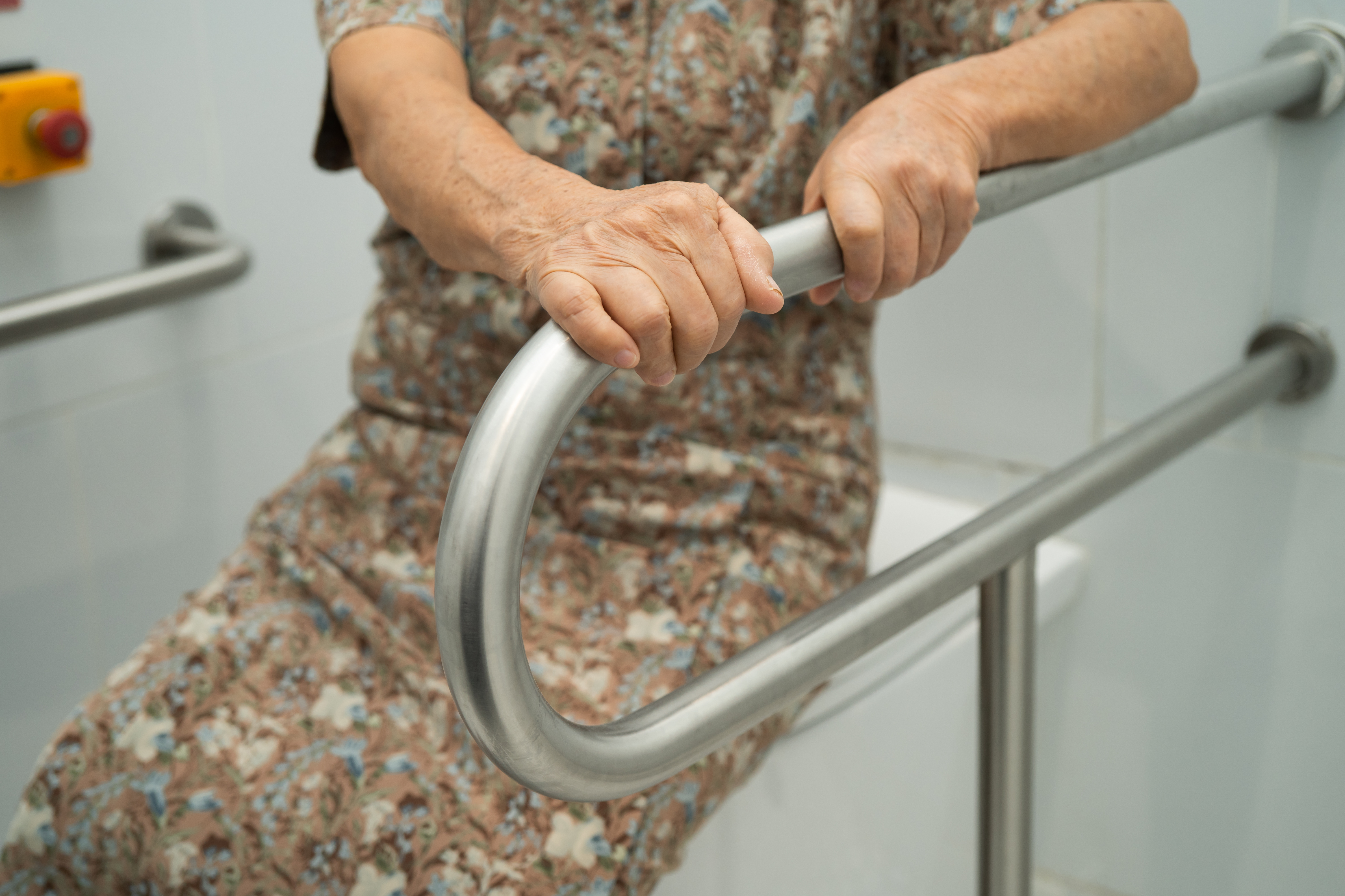 Barrierefreies Badezimmer Eine ältere Frau stzt auf einem geschlossenen Toilettendeckel, sie hält sich mit beiden Händen an einem Haltegriff fest, der neben der Toilette angebracht ist.