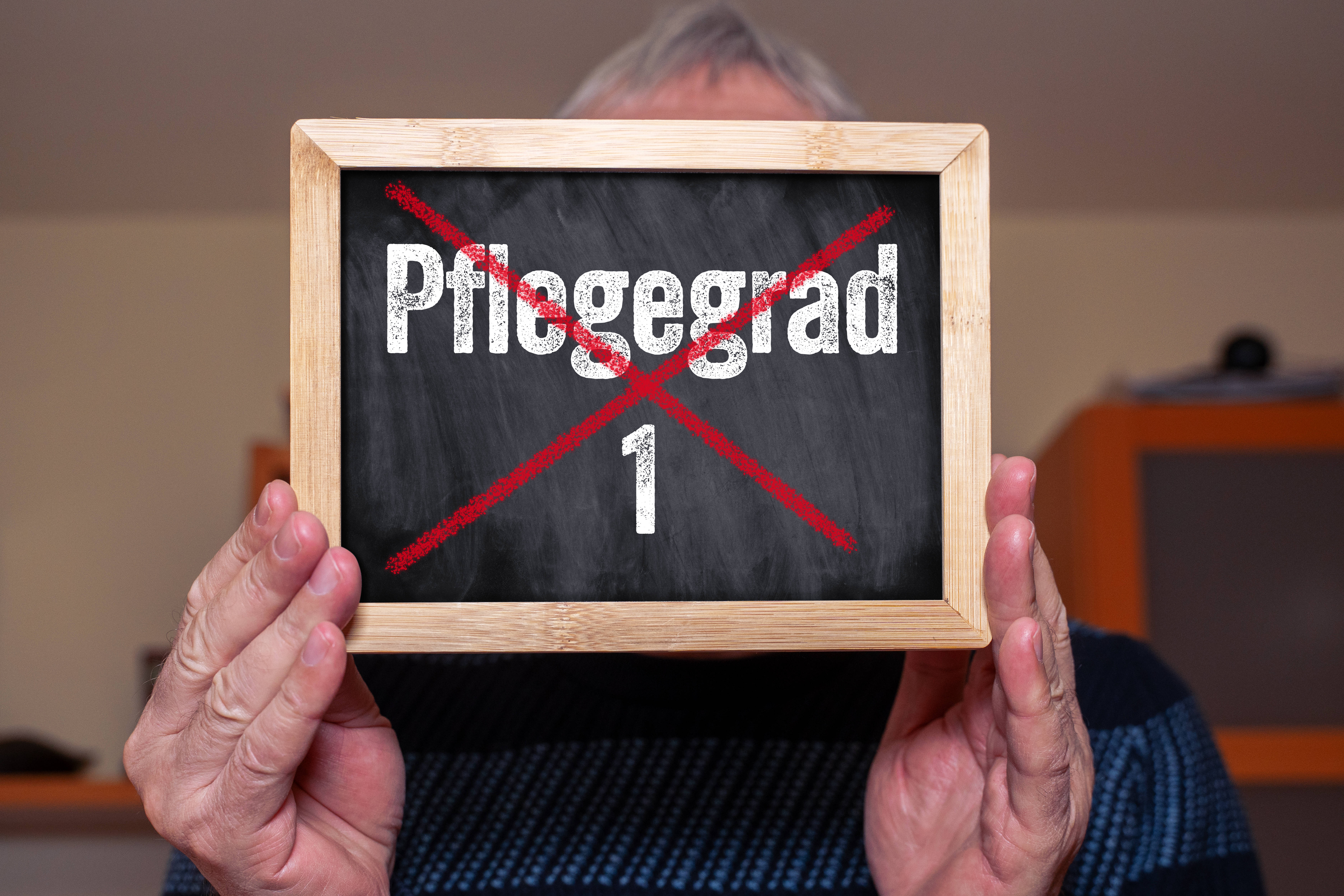 Fotomontage: Ein älterer Mann hält ein Schild mit dem durchgestrichenen Schriftzug: Pflegegrad 1. Das Symbolbild steht für die Abschaffung des Pflegegrads 1.