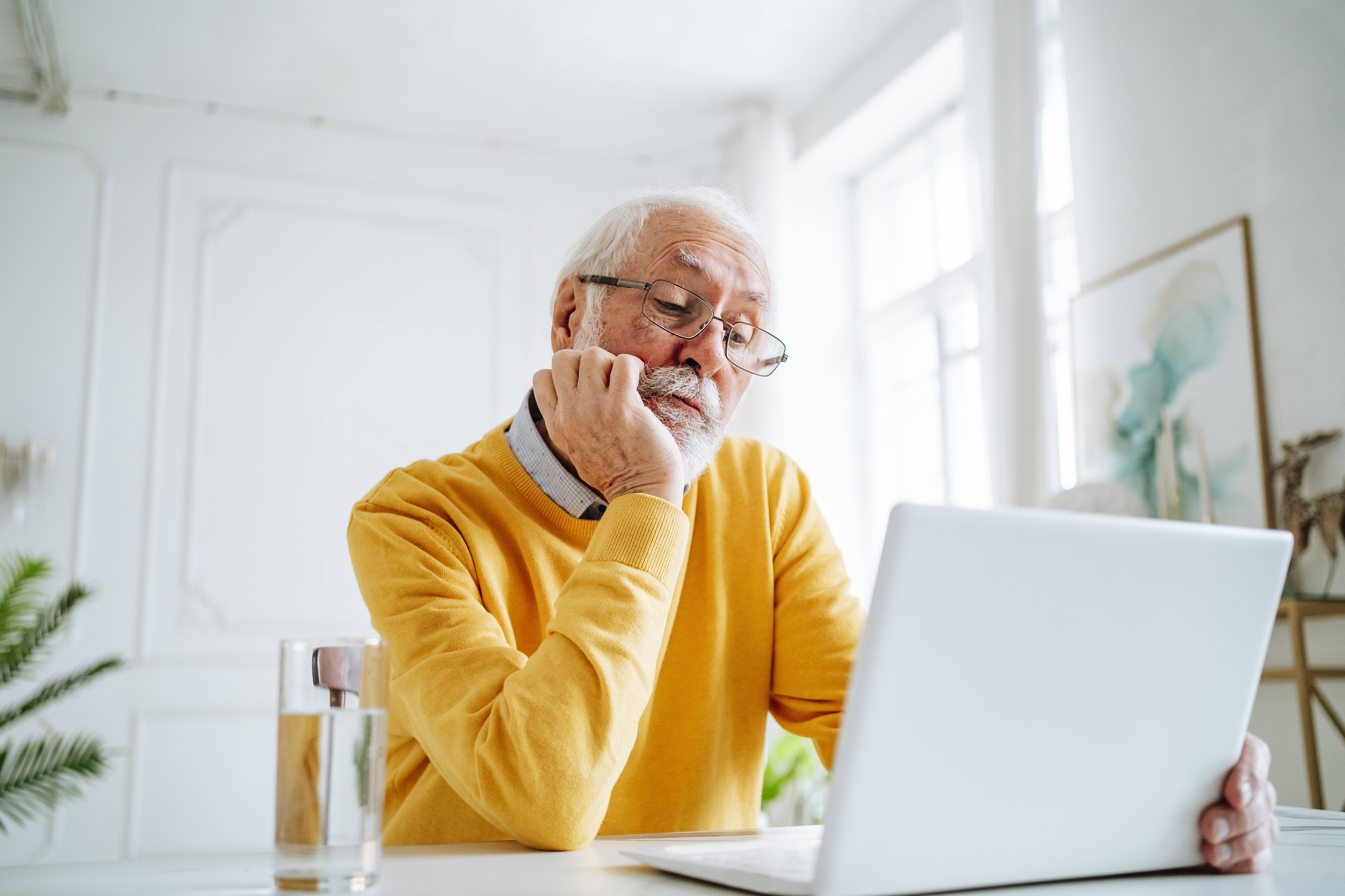 Ein Senior mit Brille im gelben Pullover sitzt an einem Tisch und schaut auf ein Notebook. Das Kinn hat er in die Hand gest&uuml;tzt.
