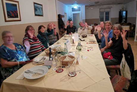 Ein paar Teilnehmer sitzen am Tisch, winken und lächeln in die Kamera