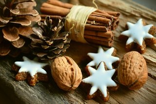 auf einem Holztisch liegen weihnachtlich geschmückt Tannenzapfen, Sternenkekse mit Zuckerglasur und Zimtstangen